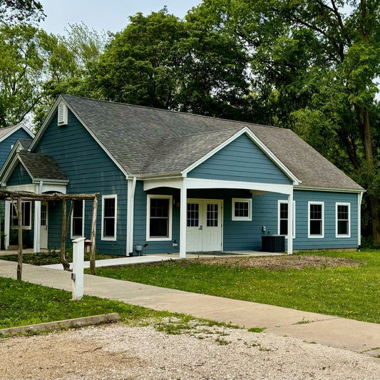Blue house with white trim surrounded by trees and a sidewalk.