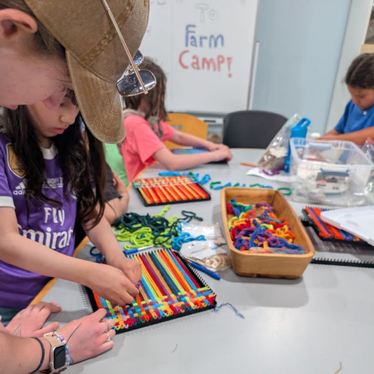 A teen wearing a tan baseball cap and purple shirt is weaving a colorful striped bracelet on a camp table, with other kids and craft supplies visible in a classroom setting.